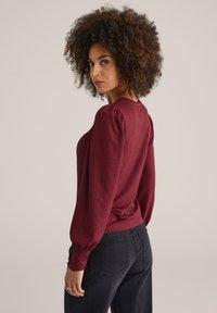 Femme aux cheveux bouclés portant un haut bordeaux à manches longues texturé et un jean noir, légèrement de côté devant un fond uni.