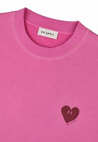 Vaaleanpunainen collegepusero, joka on valmistettu pehmeästä kankaasta. Siinä on ribbauskaulus ja sydämenmuotoinen graafinen kuvasto hymyilevällä naamalla edessä, jossa lukee "OH APRIL."