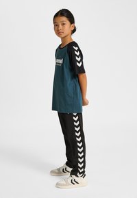 Turkos och svart T-shirt med vit text och sicksackmönster på ärmarna, kombinerad med svarta byxor med sicksackrand och ljusa sneakers.