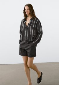 Zwart-wit gestreept shirt met kraag, losse pasvorm; gecombineerd met zwarte shorts. Model draagt zwarte instap schoenen en staat op een neutrale vloer.