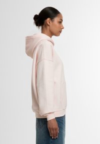 Rosa hettegenser laget av mykt stoff med frontlomme, oversized passform og ribbede mansjetter. Har snøring i hetta og avslappet silhuett.