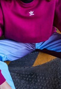 Bordowa bluza z białym logo Adidasa, w połączeniu z niebieskimi spodniami o gradiencie od jasnego do ciemnego. Materiał wydaje się gładki i miękki.