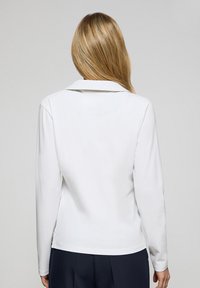 Vit långärmad topp med krage, med en jämn textur och rak fåll; visad från baksidan.