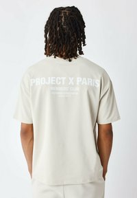 Camiseta de manga corta beige hecha de tela suave con texto gráfico blanco en la parte posterior, que presenta "PROJECT X PARIS" y "MEMBERS' CLUB."