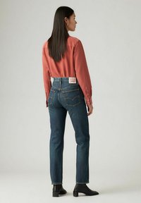 Donna con lunghi capelli scuri che indossa una camicia color ruggine, jeans blu scuro a vita alta e stivaletti neri, in piedi di spalle.