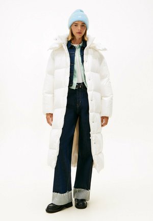 Vrouw draagt een lange witte donzen jas, een blauwe muts, een spijkerjasje, brede donkere jeans met opgerolde pijpen en zwarte schoenen, staand tegen een witte achtergrond.