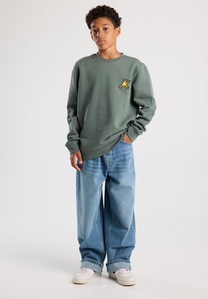Grauer Baumwoll-Sweatshirt mit einem kleinen gelben Grafik, kombiniert mit lockeren hellblauen Jeans und weißen Sneakers. Einfarbiger Hintergrund.