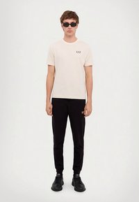 Homem vestido com uma t-shirt branca EA7, calças de jogging pretas EA7, ténis pretos e óculos de sol pretos, de pé contra um fundo branco liso.