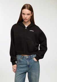 Felpa nera cropped con zip, dotata di tasca frontale e logo "Aspettami" sul lato sinistro del petto, abbinata a jeans di denim azzurri.