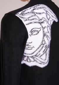 Svart kofta med intrikata vita illustrationer av ett ansikte på baksidan. Mjuk textur med detaljerat linjearbete och kontrasterande accenter.