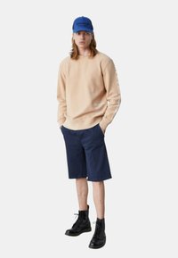 Felpa beige con scritte sulle maniche, pantaloni corti blu e stivali neri stringati. Il modello indossa un cappellino blu. Look semplice e casual.