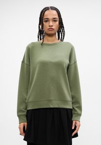 Sudadera verde oliva hecha de un tejido suave, con un corte relajado, hombros caídos y dobladillo acanalado. Llevada sobre una falda negra con pliegues.