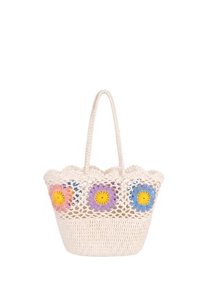 Junge Frau in braunem, geschichtetem Outfit hält eine große weiße gehäkelte Tasche mit bunten Blumenmustern und trägt beige Sandalen.