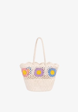 Jeune femme en tenue marron superposée, tenant un grand sac cabas blanc en crochet avec des motifs floraux colorés, portant des sandales beiges.