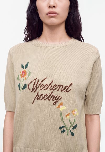 Femme portant un pull court en tricot beige avec le texte "Weekend poetry" et une broderie florale sur le devant, sur fond blanc.