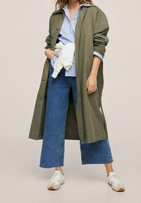 Manteau long vert olive sur une chemise rayée bleu clair, associé à un pantalon en denim à jambes larges et des baskets blanches avec des accents beiges.