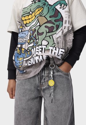Person iført grå jeans og en farverig T-shirt med en dinosaurgrafik og teksten "Mød tandlægen" samt en gul smiley-nøglering hængende i bæltestrop.