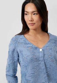 Lichtblauw doorschijnend blouse met gestructureerd bloemmotief, V-hals en een enkele witte knoop aan de voorkant. Zachte, gerafelde details overal.