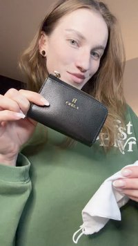 Portafoglio in pelle nera con superficie testurizzata, dettaglio logo dorato, chiusura con cerniera e forma rettangolare compatta, tenuto in una mano.