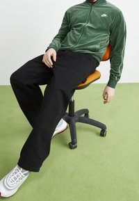 Green velvet zip-up jacket with white stripes, paired with black pants, and gray sneakers with red accents, seated on an orange chair.