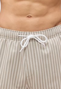 Κοντινή λήψη στο γυμνό κορμί ενός ατόμου που φοράει μπεζ και λευκό κάθετα ριγέ παντελόνι με λευκό κορδόνι δεμένο σε φιόγκο στη μέση.