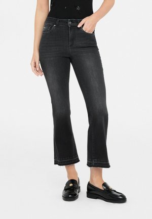 Frau trägt schwarze, hoch taillierte Cropped Flared Jeans mit ausgefransten Säumen und schwarze Loafer, Hand in der Tasche.