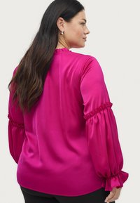 Magenta satinbluse med flæseærmer, glat tekstur og en afslappet pasform. Har en rund halsudskæring og rynkede detaljer ved manchetterne.