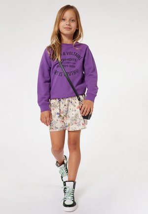 Sweat-shirt violet avec texte noir, shorts à motifs floraux et baskets montantes avec accents noir et blanc et lacets verts. Sac bandoulière noir.
