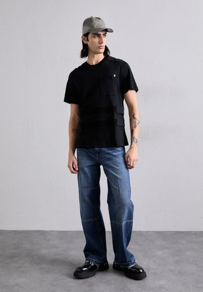 T-shirt nera a maniche corte con tasca sul petto e bande sovrapposte, abbinata a jeans blu a vestibilità ampia e scarpe chunky nere. Cappellino grigio indossato.
