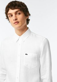 Camicia bianca a maniche lunghe in lino con colletto con bottoni, dotata di una tasca sul petto e un piccolo logo ricamato nero.