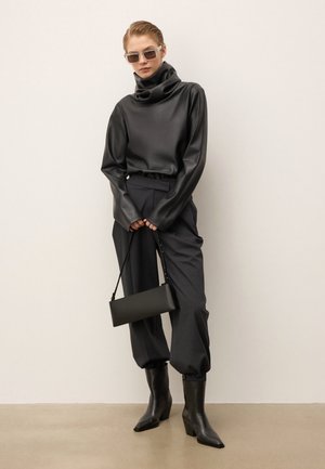 Mujer vistiendo una blusa negra de cuero de cuello alto, pantalones oscuros ajustados, botas negras de tacón, gafas de sol y sosteniendo un bolso rectangular negro.