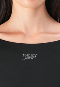 Černý top s vykrojeným výstřihem a stříbrnou výšivkou "tommy jeans" na hrudi. Hladký materiál s přiléhavým střihem.