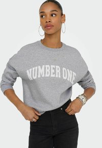 Sudadera gris con el texto "NÚMERO UNO" en un diseño arqueado en blanco. Presenta puños acanalados y cuello redondo. Combinada con jeans negros.