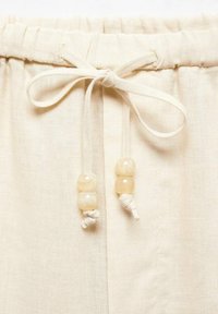 Lněná šňůrka světle krémové barvy, na jejích koncích dvě kulaté, světlé korálky. Texturovaná tkanina.