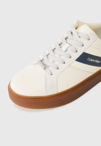 Fehér sportcipő texturált felülettel, szürke fűzőkkel és barna gumitalppal. Navy csík jellemzi, amelyen a "Calvin Klein" logó található.