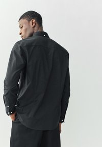 Camisa negra de manga larga con cuello abotonado, textura suave y puños abotonados. Corte entallado con bajo curvado.