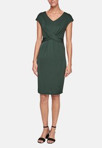 Grön klänning i mjukt tyg med V-ringning och korta ärmar. Har en tvistad framdesign och är knä-lång. Svarta remsandaler.