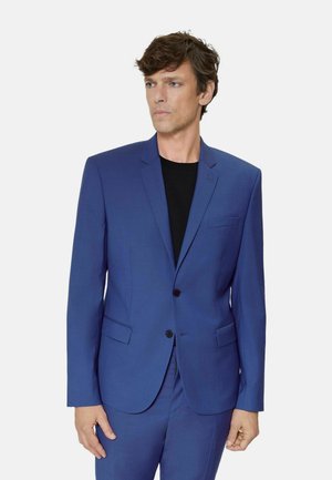 Uomo che indossa un giacca di abito blu su misura e pantaloni abbinati sopra una camicia nera, in piedi davanti a uno sfondo chiaro uniforme.