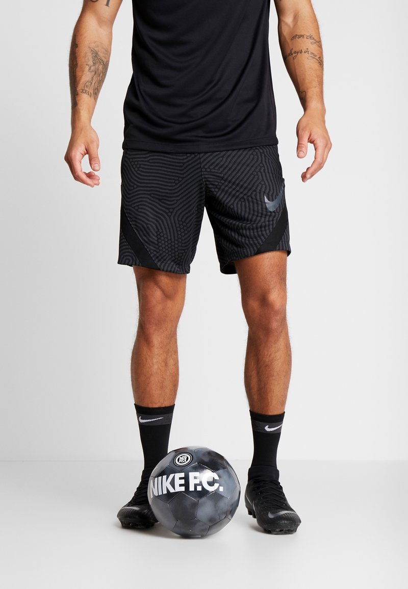 Mężczyzna w czarnych, wzorzystych szortach i skarpetkach stoi z jedną stopą obok czarno-szarej piłki do piłki nożnej Nike F.C. na białej podłodze.