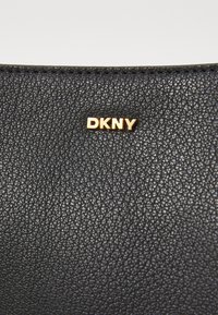 Texture in pelle nera con una sottile venatura, caratterizzata da un accento del logo DKNY in oro sulla superficie. Focus sul materiale e sui dettagli.