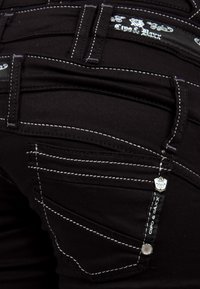 Zwarte jeans met witte stiksels, voorzien van meerdere zakken en een decoratief label. Materiaal is denim met een gladde textuur.