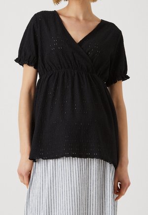 Vrouw draagt een zwarte blouse van oogjesstof met pofmouwen en een V-hals over een wit-grijs gestreepte rok.