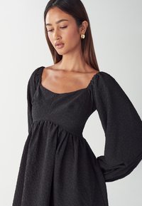 Schwarzes Kleid aus strukturiertem Stoff, mit quadratischem Ausschnitt und puffierten langen Ärmeln. Der flared Rock sammelt sich an der Taille und erzeugt eine fließende Silhouette.