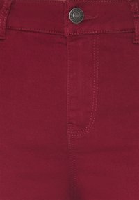 Primer plano de una tela de mezclilla roja que muestra un botón, un lazo para el cinturón y costuras detalladas alrededor del área de la cremallera.