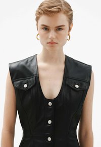 Vestido sin mangas de cuero negro con un escote en V profundo, dos bolsillos en el pecho con broches y seis botones en la parte delantera. Textura suave y corte entallado.