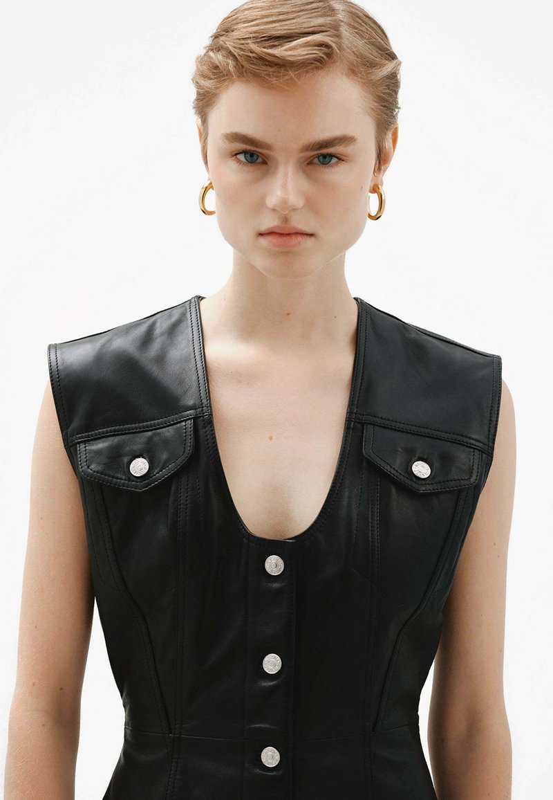 Vestido sin mangas de cuero negro con un escote en V profundo, dos bolsillos en el pecho con broches y seis botones en la parte delantera. Textura suave y corte entallado.