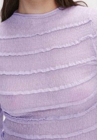 Gros plan sur une personne portant un haut lavande transparent avec des rayures horizontales à volants et un col rond.