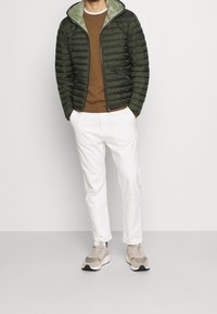 Grön olivfärgad puffjacka för män med en kontrasterande insida, kombinerad med en brun tröja, vita byxor och beige sneakers.