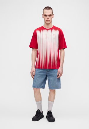Jeune homme debout face à l’avant, portant un T-shirt tie-dye rouge et blanc, un short en jean bleu, des chaussettes blanches et des baskets noires.
