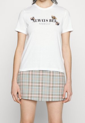 Kobieta w białym T-shircie z pszczołami i tekstem "Always Bee Yourself", zestawionym z pastelową plisowaną mini spódnicą.